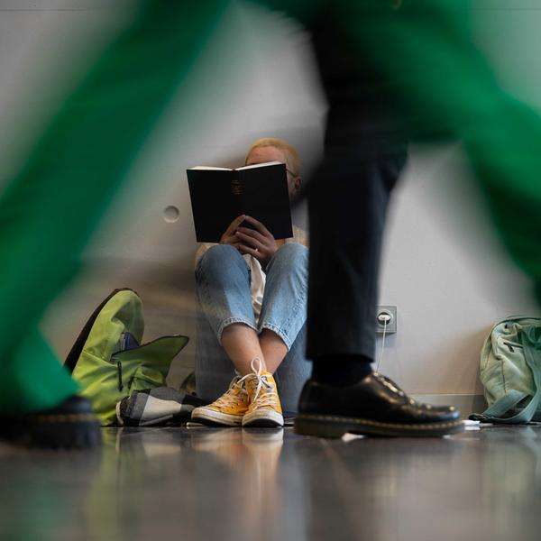 Eine Person sitzt während der Frankfurter Buchmesse lesend an einer Wand, während im Vordergrund Messebesuchende vorbei laufen.