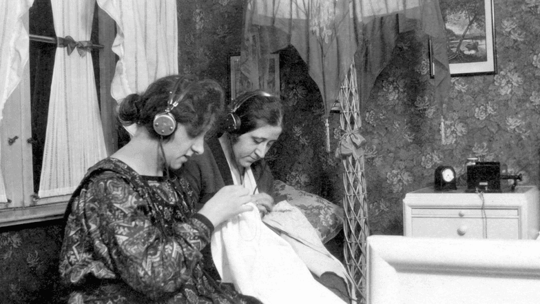 Zwei Frauen sitzen über Stickarbeiten und hören dabei Radio über Kopfhörer.