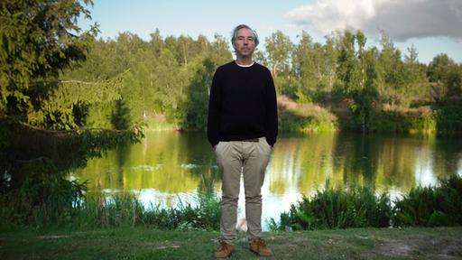 Olli Schulz steht am grasbewachsenen Ufer eines Teichs.