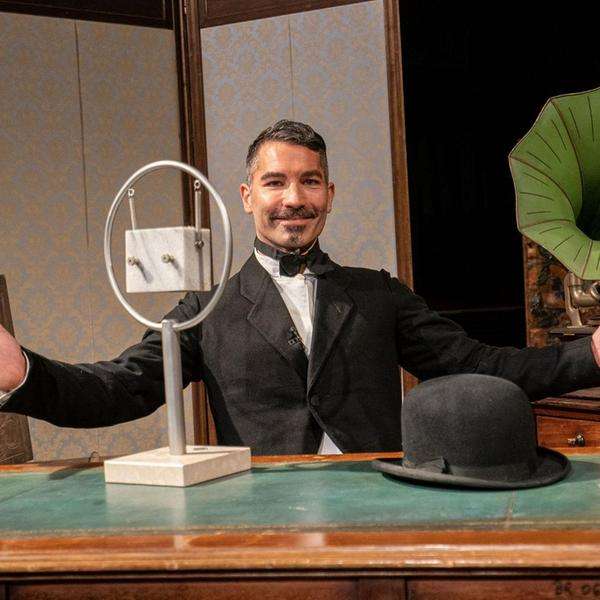 Ein Mann im Anzug sitzt mit weit ausholender Geste vor Theaterrequisiten: Grammophon und altes Radiomikrofon, ein Hut liegt auf dem Tisch vor ihm