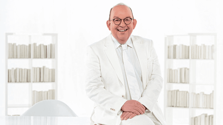 Denis Scheck, ein Mann, mittleren Alters mit Brille und Halbglatze sitzt in weißem Anzug auf einem Tisch.