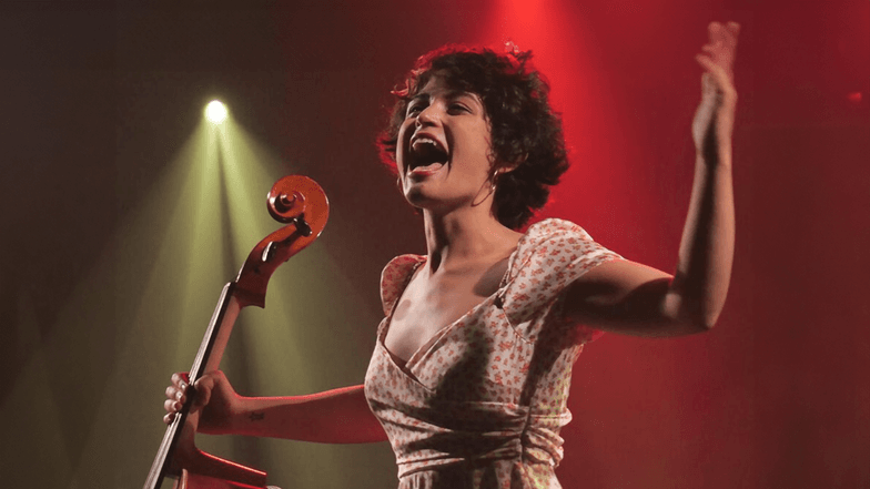Eine junge Jazz-Sängerin mit einem Cello in der rechten Hand steht auf einer Bühne.