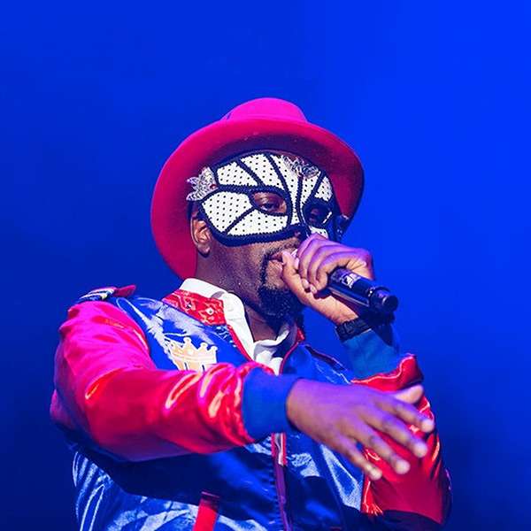 Wyclef Jean auf der Bühne mit Maske, rotem Hut und blauer Jacke.